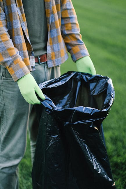 A person wearing a yellow and grey checkered shirt, with a green shirt underneath, and grey trousers, is holding open a large black plastic rubbish bag outdoors on a grassy area. The individual is wearing light green gloves and appears to be preparing to dispose of waste or rubbish. The background shows a well-maintained lawn, suggesting a garden or park setting. The scene reflects typical waste collection activities, possibly related to private rubbish removal services like those provided by Rubbish Clearance Dulwich, emphasizing the importance of proper waste handling outside of local authority collections, and highlighting on-site rubbish disposal practices.