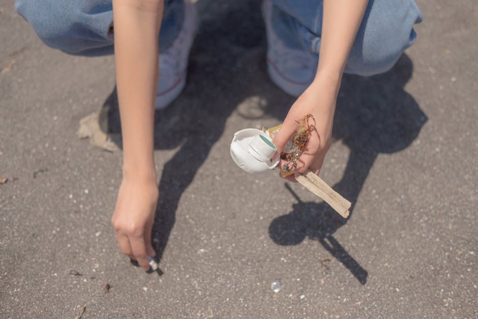 A person wearing light blue jeans and white sneakers is crouching on a paved surface, holding a small wooden stick with a damaged, partially broken white plastic or ceramic object attached to it. The object appears to be broken and is interwoven with some fibrous, reddish-brown material. The background shows the paved ground with small debris scattered around. The scene suggests disposal or collection of small waste items, aligning with the context of rubbish removal services offered by Rubbish Clearance Dulwich, as seen on the webpage about East Dulwich Lordship Lane rubbish removal tips. The lighting indicates natural daylight, and the environment appears outdoor, possibly on a driveway or street area dedicated to waste collection or clearance activities.