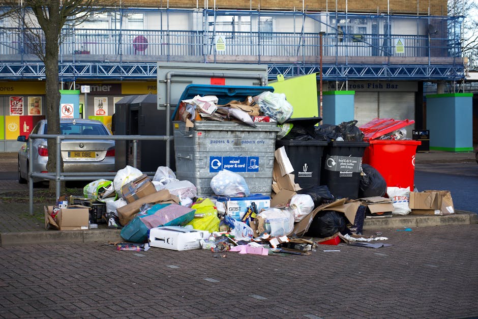 A large pile of mixed household waste and rubbish surrounds a central recycling bin labeled for paper and cardboard, located on a paved urban street near parked cars. The debris includes crumpled paper, cardboard boxes, plastic bags, and packaging materials, some spilling onto the sidewalk and curb. To the right of the main bin are additional black and red rubbish bins, partially filled with waste, with black plastic trash bags also visible nearby. Behind the waste collection area, a storefront with signage and a shopfront with closed shutters can be seen, along with a building under construction or maintenance, covered in scaffolding and blue netting. The scene appears to be in a residential or commercial area, and the overall environment suggests the need for timely rubbish collection, potentially handled by private waste removal services like Rubbish Clearance Dulwich. The image emphasizes the importance of regular rubbish disposal and alternative waste handling methods to maintain urban cleanliness.