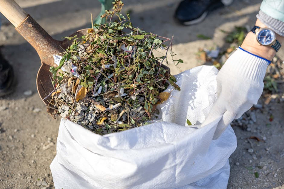 A person wearing a white glove and a wristwatch is holding a white reusable bag filled with garden waste, including small branches, leaves, and plant stems, near the ground. The bag appears to be partially open, revealing the debris inside. There is a rusty hand tool, possibly a shovel, resting on or near the bag, with its blade covered in dirt and soil. The ground surface is a mix of soil and small gravel, with some scattered leaves and bits of debris around. In the background, part of a person's leg and shoe are visible, indicating an outdoor setting. This scene is consistent with a rubbish removal or garden clearance activity, often handled by private waste disposal services like Rubbish Clearance Dulwich, supporting alternative waste handling methods such as on-site clearance or private collection of garden waste.