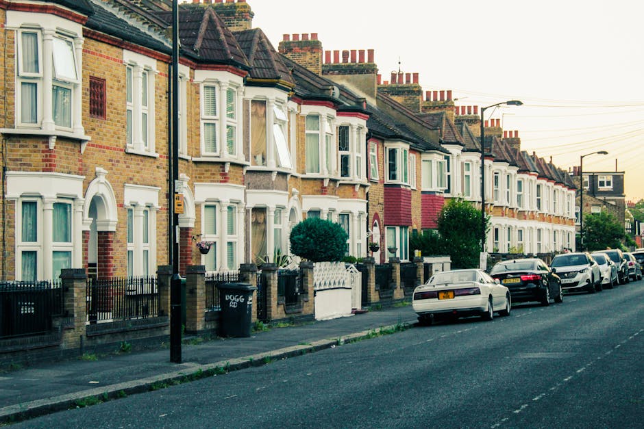 A row of traditional terraced houses on a residential street featuring brick facades with decorative white window surrounds and bay windows, some with open or partially open sash windows revealing curtains inside. The houses have pitched tiled roofs with multiple red-brick chimney stacks, and each property is separated by low brick walls topped with wrought iron fencing. Small front gardens with neatly trimmed bushes and plants are visible in front of several homes. Parked along the street are various cars, including sedans and hatchbacks in dark, light, and metallic tones, parked parallel to the curb on the right side of the image. The pavement is paved with concrete slabs, and street lamps are positioned intermittently along the sidewalk, with some extending over the road. The sky appears clear with soft evening light, casting gentle shadows on the buildings. The scene suggests a typical UK residential area, possibly near a property management or private waste disposal service such as Rubbish Clearance Dulwich, with no visible waste or rubbish in the scene itself but highlighting the tidy, well-maintained environment typical of private clearance or rubbish removal settings.