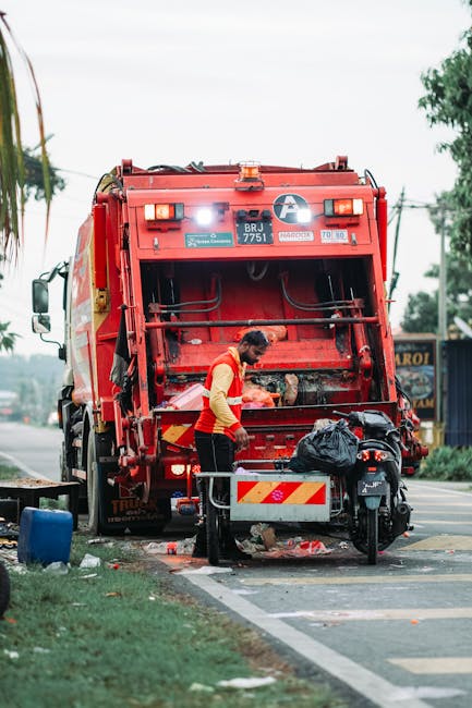 A waste collection worker wearing a high-visibility orange and yellow jacket is operating a large red rubbish collection truck on the side of a paved street. The truck's rear hopper is open, revealing mechanical components and a dark, textured interior designed for waste intake. The worker is leaning slightly forward, inspecting or managing waste disposal, with a black garbage bag placed nearby on a small stand attached to the truck. The surrounding environment includes a patch of green grass at the edge of the pavement, a blue container on the ground, and distant trees and power lines under a cloudy sky. The scene depicts an outdoor waste collection operation, consistent with private refuse handling services, and the vehicle and worker are positioned close to a curb, indicating a typical urban or suburban roadside setting. The image emphasizes the practical aspects of rubbish removal, demonstrating the machinery and personnel involved in efficient waste management outside of standard municipal collection.