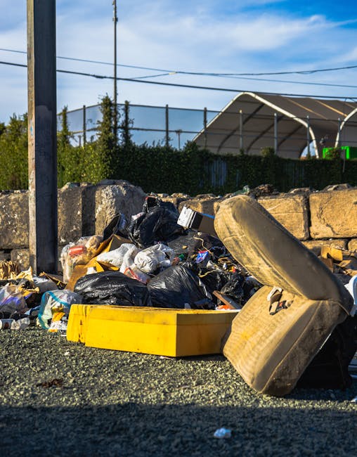 A large accumulation of mixed waste materials is visible in an outdoor area, consisting of black plastic refuse bags, scattered debris, and a yellow plastic container lying on its side. There is an old, worn car tire leaning against the pile and a stone wall made of irregularly shaped rocks in the background. The scene is set on a gravel surface with some small pieces of rubbish spread around. Behind the waste, there is a metal fence and some greenery, with a partly cloudy sky overhead. The environment suggests a temporary rubbish collection point, possibly part of an independent waste clearance service, with the clutter indicating rubbish removal or on-site clearance activity managed by Rubbish Clearance Dulwich or similar providers.
