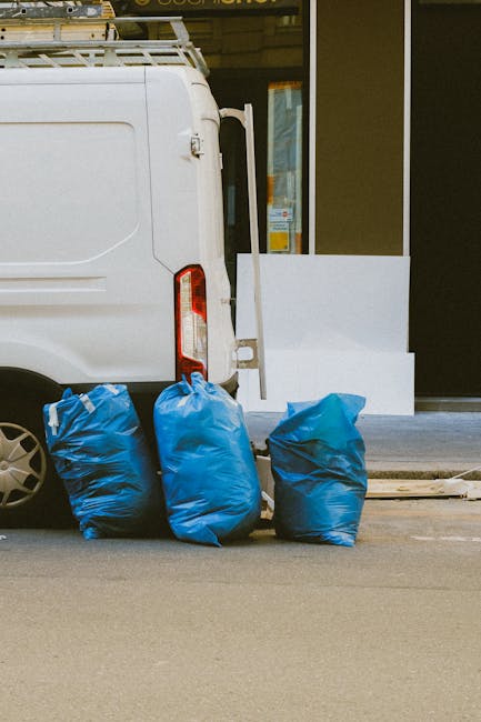 A waste collection worker wearing a high-visibility orange and yellow jacket is operating a large red rubbish collection truck on the side of a paved street. The truck's rear hopper is open, revealing mechanical components and a dark, textured interior designed for waste intake. The worker is leaning slightly forward, inspecting or managing waste disposal, with a black garbage bag placed nearby on a small stand attached to the truck. The surrounding environment includes a patch of green grass at the edge of the pavement, a blue container on the ground, and distant trees and power lines under a cloudy sky. The scene depicts an outdoor waste collection operation, consistent with private refuse handling services, and the vehicle and worker are positioned close to a curb, indicating a typical urban or suburban roadside setting. The image emphasizes the practical aspects of rubbish removal, demonstrating the machinery and personnel involved in efficient waste management outside of standard municipal collection.